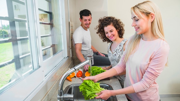 Szczęśliwa para patrzeje młodego żeńskiego przyjaciela myje sałaty w kuchennym zlew