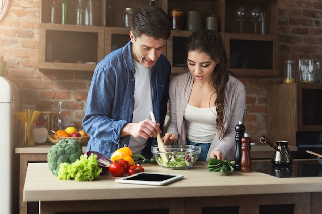 Szczęśliwa para przygotowuje kolację z przepisem w tablecie w swojej kuchni na poddaszu w domu. Przygotowanie sałatki warzywnej.