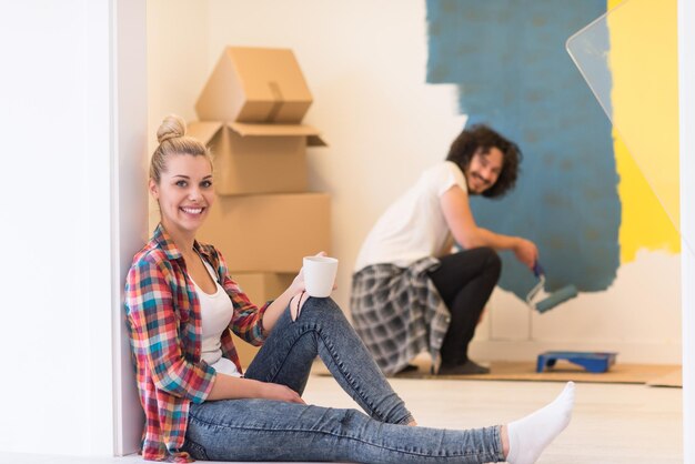 Szczęśliwa para robi remont domu, mężczyzna maluje pokój, a kobieta relaksuje się na podłodze i pije kawę