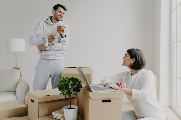 Szczęśliwa para rodzinna wprowadza się do nowego mieszkania Żona pracuje na laptopie wyszukuje informacje w internecie sprawia, że zakupy online mężczyzna stoi z psem wypakowuje rzeczy z pudeł