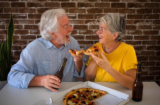 Szczęśliwa para seniorów bawi się razem jedząc pizzę Starsza kobieta i mężczyzna śmieją się, siedząc w domu przy stole z piwem i pizzą