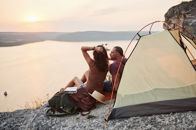 szczęśliwa para siedzi w namiocie z widokiem na jezioro podczas wycieczki pieszej. koncepcja wakacji przygodowych