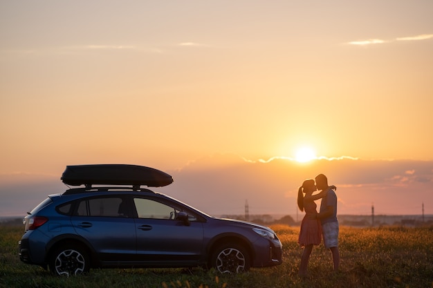 Szczęśliwa para spędza razem czas w pobliżu ich samochodu SUV podczas podróży poślubnej w ciepły letni wieczór. Młody mężczyzna i kobieta korzystających z podróży samochodem w przyrodzie.