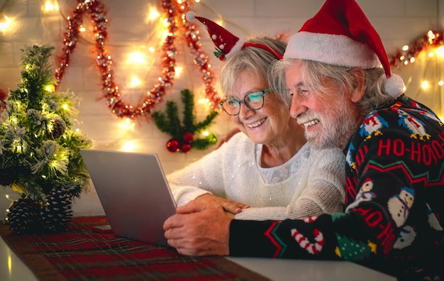 Szczęśliwa para starszych w kapeluszu Świętego Mikołaja w czasie świąt Bożego Narodzenia podczas rozmowy wideo przez laptop z domu, kontaktując się z rodziną lub przyjaciółmi