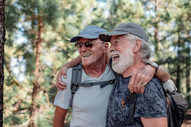 Szczęśliwa para starych aktywnych mężczyzn w kapeluszu i plecaku obejmuje się i bawi w górskiej wędrówce po lesie, doceniając przygodę i wolność emerytowanych seniorów i koncepcję zdrowego stylu życia