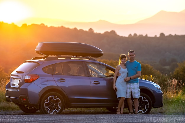 Szczęśliwa para stoi w pobliżu ich samochodu o zachodzie słońca. Młody mężczyzna i kobieta, ciesząc się razem, podróżując pojazdem.