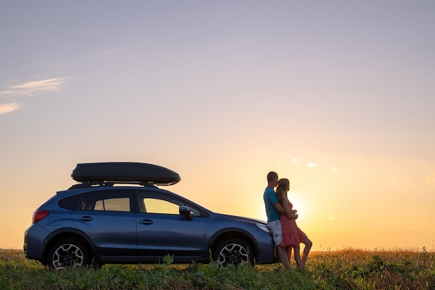 Szczęśliwa para stojąca obok swojego samochodu o zachodzie słońca Młody mężczyzna i kobieta cieszą się wspólnym czasem podróżując pojazdem
