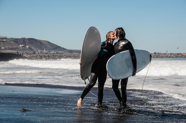 Zdjęcie szczęśliwa para surferów w czarnych kombinezonach trzyma białą deskę i całuje w wodzie na tle nieba i oceanu