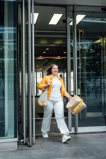 Szczęśliwa pewna siebie uśmiechnięta plus size kręta młoda kobieta z torbami na zakupy i modną siatkową torbą spacerującą po ulicy miasta w pobliżu okien centrum handlowego