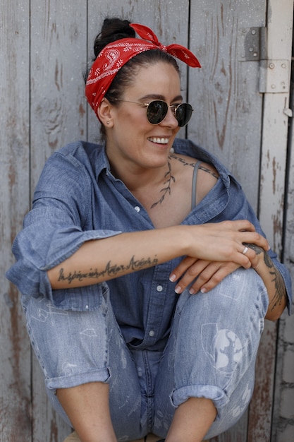 Szczęśliwa piękna Amerykanka ze stylowymi okularami przeciwsłonecznymi w dżinsowej koszuli i dżinsach siedzi w pobliżu drewnianej ściany. Styl pinup