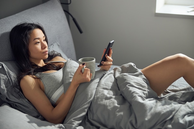 Szczęśliwa piękna azjatykcia kobieta relaksuje się w łóżku podczas korzystania ze smartfona w godzinach porannych