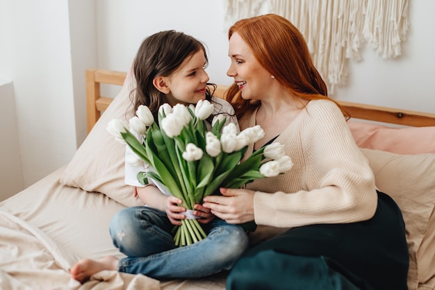 Szczęśliwa piękna czuła mama z bukietem wiosennych kwiatów przytula córkę w domu na łóżku cieszy się chwilą niespodzianki świętuje Dzień Matki