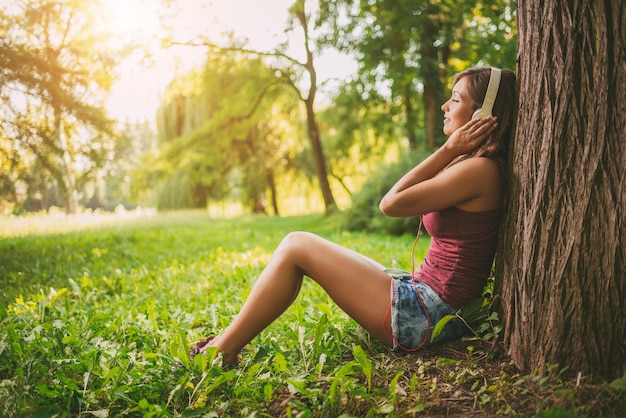 Szczęśliwa piękna dziewczyna siedzi przy drzewie w parku i słuchając muzyki.