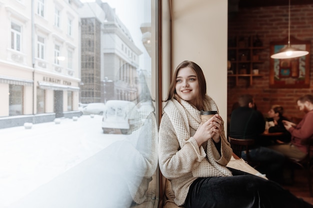 Szczęśliwa piękna kobieta w białym swetrze i szaliku siedzi w pobliżu okna w kawiarni