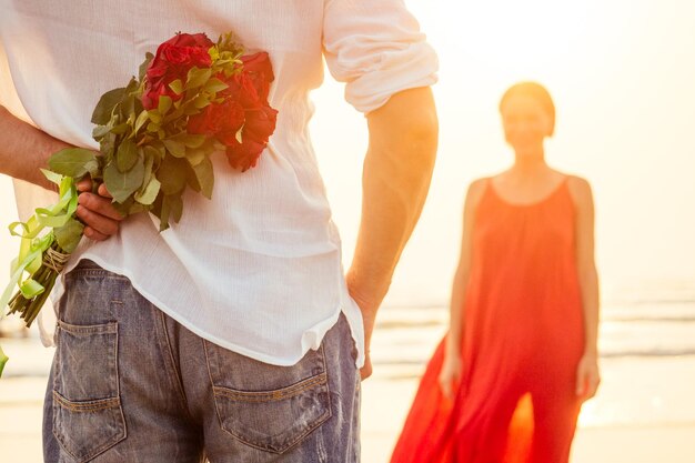 Szczęśliwa piękna kobieta w czerwonej sukience odbierająca prezent od mężczyzny nad morzem zachód słońcaniespodzianka żona od męża bukiet róż dzień kobiet 8 marca i walentynki zaręczyny miłość od pierwszego wejrzenia romantyczna