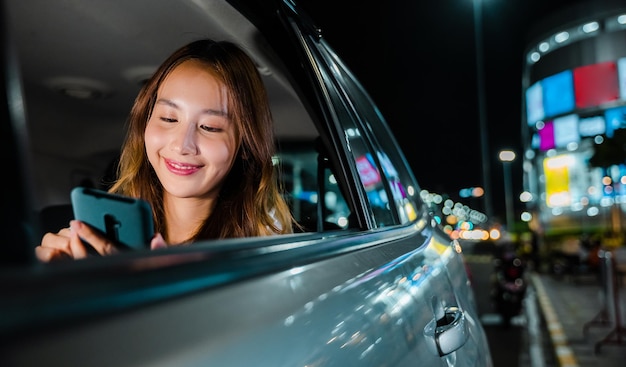 Szczęśliwa piękna kobieta wysyłająca SMS-y do smartfona siedzącego na tylnym siedzeniu samochodu w mieście, azjatycka bizneswoman dojeżdżająca z biura na tylnym siedzeniu taksówki z telefonem komórkowym na drodze w mieście w nocy po późnej pracy