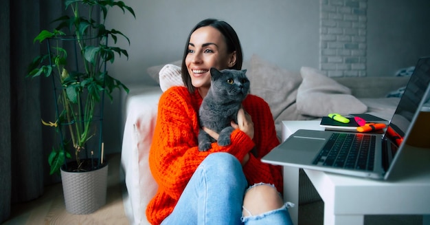 Szczęśliwa piękna młoda brunetka trzyma i przytula swojego słodkiego brytyjskiego kota krótkowłosego na rękach po pracy na laptopie w domu