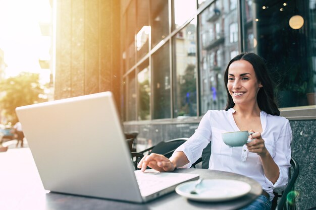Szczęśliwa piękna młoda kobieta pracuje na laptopie w ulicznej kawiarni na świeżym powietrzu
