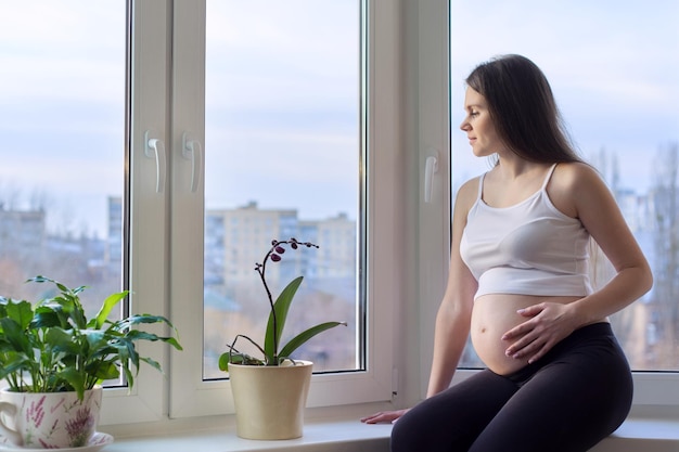 Szczęśliwa piękna młoda kobieta w ciąży marzycielsko patrząc przez okno domu