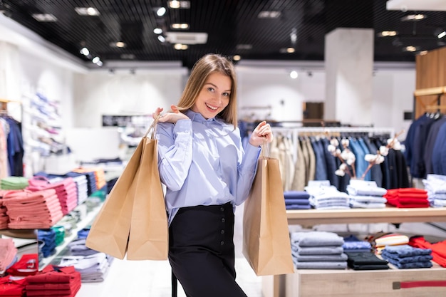 Szczęśliwa piękna młoda kobieta z modnymi efektownymi ubraniami z papierowymi torbami w sklepie