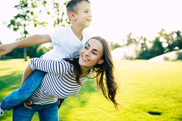 Szczęśliwa piękna młoda mama bawi się ze swoim synkiem, gdy jedzie na jej plecach i krzyczy z radości