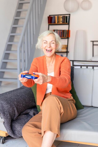 Zdjęcie szczęśliwa piękna starsza szaro włosy starsza kobieta siedząca na kanapie w salonie przytulne mieszkanie uśmiechnięta starsza kobiety z zdalnym sterowaniem do gry konsolę nowoczesna babcia gra w gry wideo online