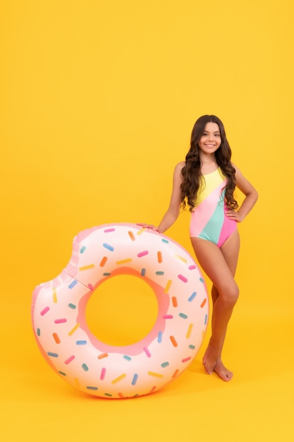 szczęśliwa plaża nastolatka z kręconymi włosami w stroju kąpielowym z nadmuchiwanym pierścieniem pączka do zabawy na basenie na letnie wakacje na żółtym tle, akcesoria plażowe.