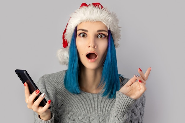 Szczęśliwa Podekscytowana Młoda Kobieta W Santa Hat Z Smartphone Na Szarym Tle. Boże Narodzenie Koncepcja Sprzedaży Zakupów Online