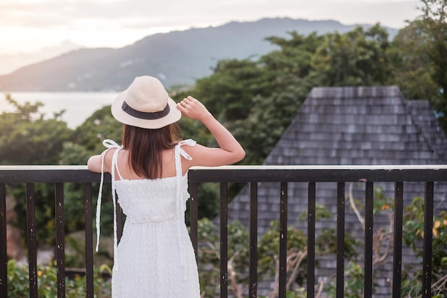 Szczęśliwa podróżniczka kobieta w białej sukni i kapeluszu cieszyć się pięknym widokiem na morze młoda kobieta stojąca na tropikalnej plaży Wolność relaksujące wakacje wakacje i koncepcja podróży letnich
