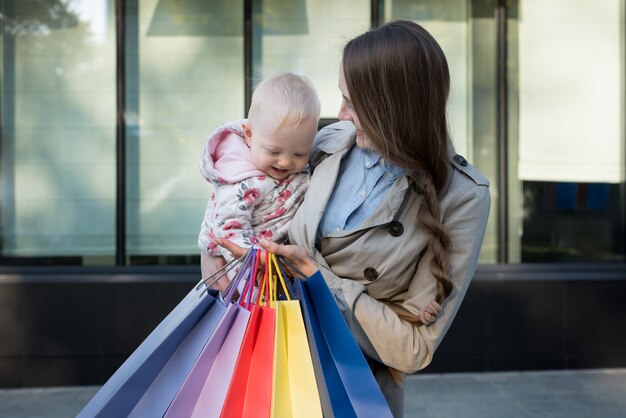 Zdjęcie szczęśliwa potomstwo matka z małą córką na rękach i torba na zakupy w ręce. dzień zakupów. centrum handlowe
