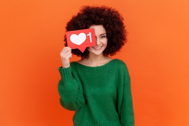 Szczęśliwa pozytywna młoda dorosła kobieta z fryzurą afro na sobie zielony sweter w stylu casual, zakrywający jej oko z sercem w mediach społecznościowych, jak ikona. Kryty studio strzał na białym tle na pomarańczowym tle.