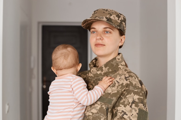Zdjęcie szczęśliwa pozytywna optymistyczna dumna młoda dorosła kobieta żołnierz ubrana w mundur kamuflażowy, wracająca do domu i zabierająca swoją małą córeczkę, odwracając wzrok z zadowolonym wyrazem twarzy.