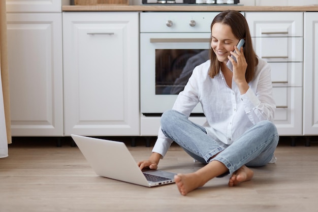 Szczęśliwa pozytywna optymistyczna kobieta ubrana w białą koszulkę i dżinsy, siedząc na podłodze w kuchni i za pomocą laptopa do pracy, mając przyjemną rozmowę przez inteligentny telefon.