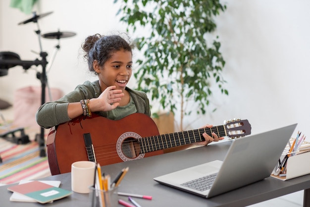 Szczęśliwa preteen uczennica z gitarą macha ręką do swojego nauczyciela na ekranie laptopa podczas lekcji muzyki online