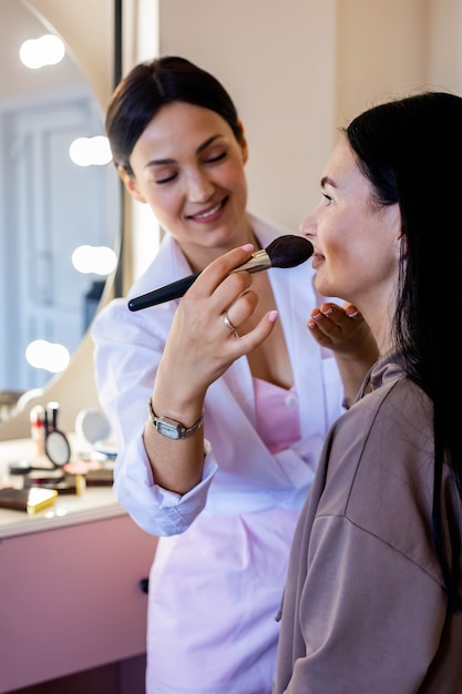 Szczęśliwa profesjonalna wizażystka nakładająca kosmetyki kredkę do ust klientce pracującej w salonie kosmetycznym