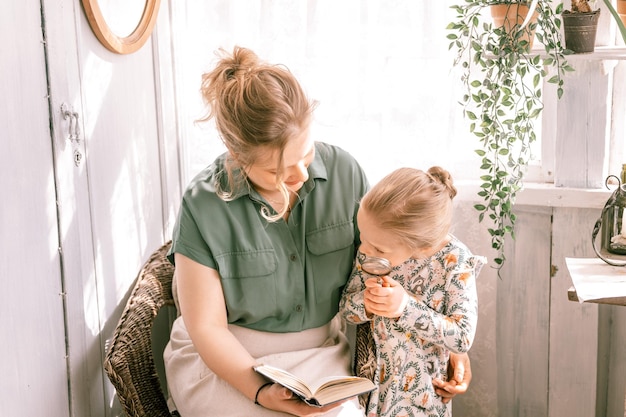 Szczęśliwa rodzina matka z uroczą małą córką córką bawią się razem na przedmieściach wiejskiego domu w słoneczny letni dzień szczera mama przytula się i czyta książkę dziecku odkrywając książkę z lupą