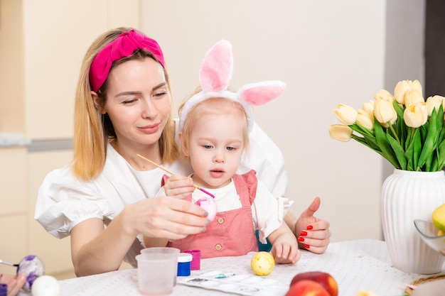 Zdjęcie szczęśliwa rodzina przygotowuje się do wielkanocy śliczna dziewczyna z matką maluje jajka aktywność domowa pojęcie jedności i miłości mama i córka