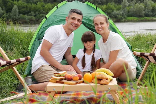 Szczęśliwa rodzina siedząca w pobliżu namiotu na ziemi podczas pikniku, ciesząc się smacznymi owocami i kanapkami, spędzając szczęśliwy weekend na świeżym powietrzu