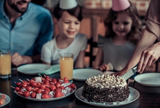 Zdjęcie szczęśliwa rodzina siedzi przy stole w udekorowanej kuchni podczas urodzin wykadrowany obraz mamy krojącej tort urodzinowy