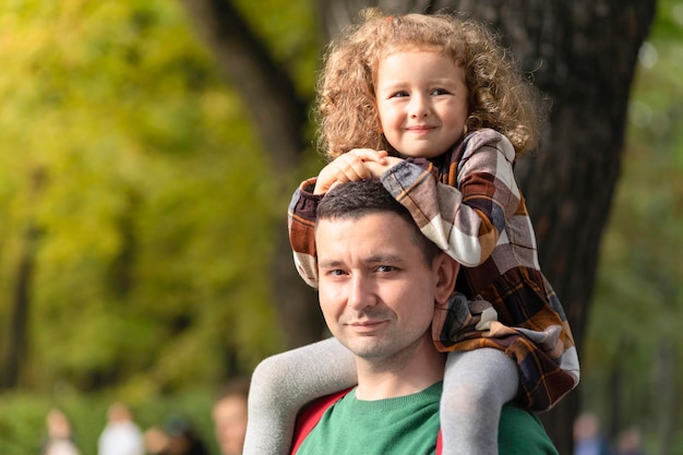 Szczęśliwa rodzina. śmiejąc się córka siedzi na ramionach taty. mała dziewczynka z ojcem w parku. uśmiechnięty mężczyzna niosący dziecko