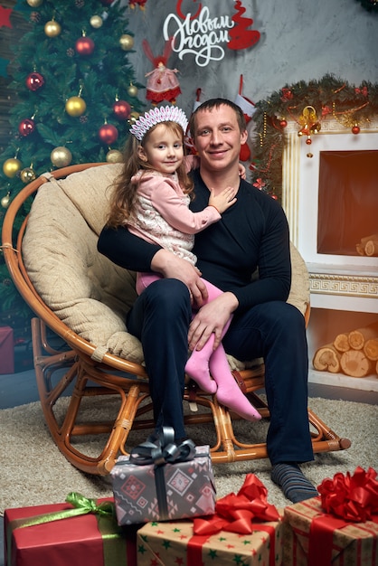 Szczęśliwa rodzina spędza razem czas na ferii zimowych w domu przy kominku przy choince z prezentami. Śliczna mała dziewczynka z ojcem na krześle przy choince.