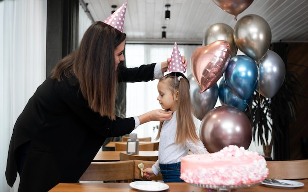 Szczęśliwa rodzina świętująca urodziny lub rocznicę mama ubiera córkę w imprezową czapkę Tort urodzinowy i balony