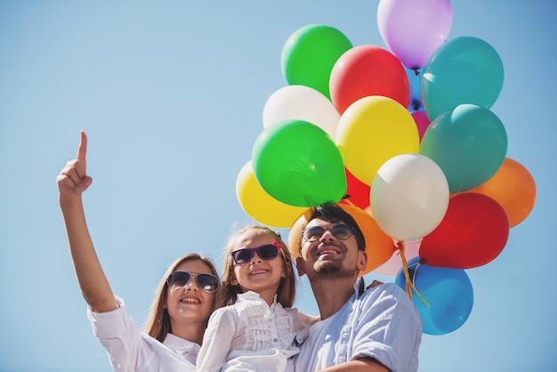 Szczęśliwa rodzina z balonami patrzącymi na zewnątrz w letni dzień, mama wskazująca na górę.