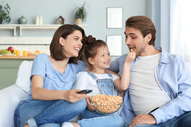 Szczęśliwa rodzina z dzieckiem siedzi na kanapie oglądając telewizję i jedząc popcorn, młodzi rodzice obejmując córkę razem relaks na kanapie.