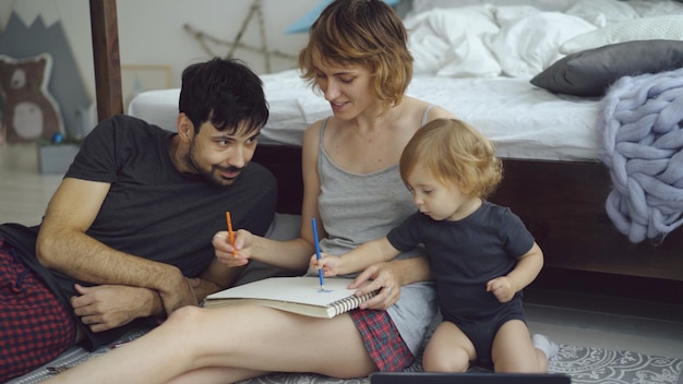 Szczęśliwa rodzina z uroczą uroczą córką rysującą w albumie ołówkami siedzącą w domu w pobliżu łóżka