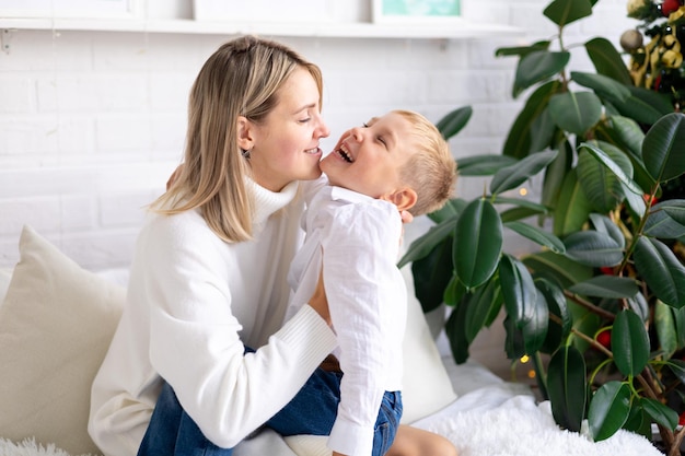 Szczęśliwa rodzinna mama i syn w białych swetrach i dżinsach na łóżku w jasnej sypialni w domu przytulają się i uśmiechają