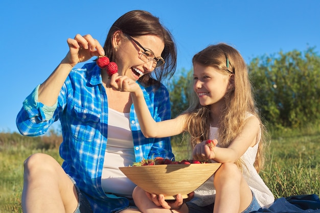 Szczęśliwa roześmiana matka i córka dziecka razem na zewnątrz, siedząc na zielonej trawie, piknik z miską świeżych truskawek. Dzień Matki, majowa natura