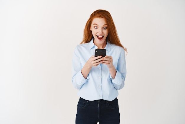 Szczęśliwa Ruda Kobieta Krzyczy Z Radości I Zaskoczenia, Czytając Ofertę Promocyjną Na Ekranie Smartfona, Uśmiechając Się Zdumiona Stojąc Na Białym Tle