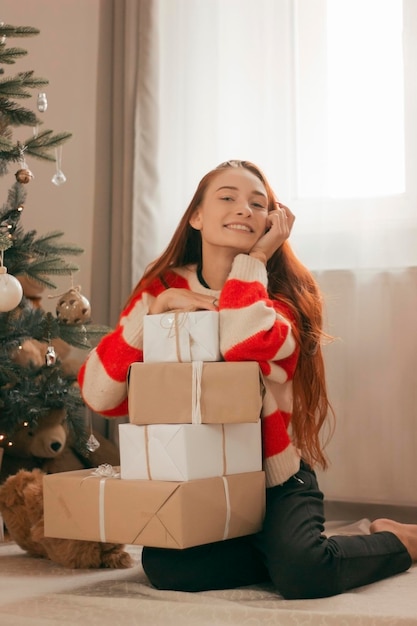 Szczęśliwa ruda kobieta w swetrze z prezentami pod choinką ze światłami święta noworoczne