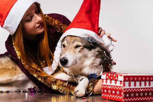 Szczęśliwa rudowłosa dziewczyna w czerwonej czapce jak Święty Mikołaj siedzi na podłodze z psem, noworoczna atmosfera, złoty blichtr i prezenty